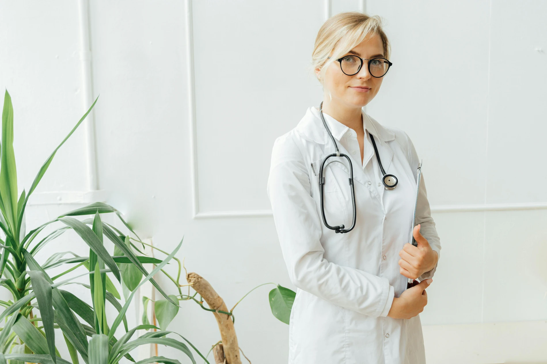 A smiling blonde woman in a white lab coat and glasses holds a clipboard. She’s a panel physician for immigration exams.