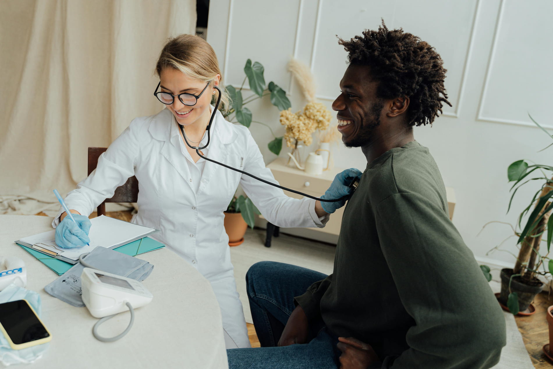 Immigration Medical Exam (IME) preparation. A doctor listens to a smiling patient's chest with a stethoscope while taking notes for an IME.