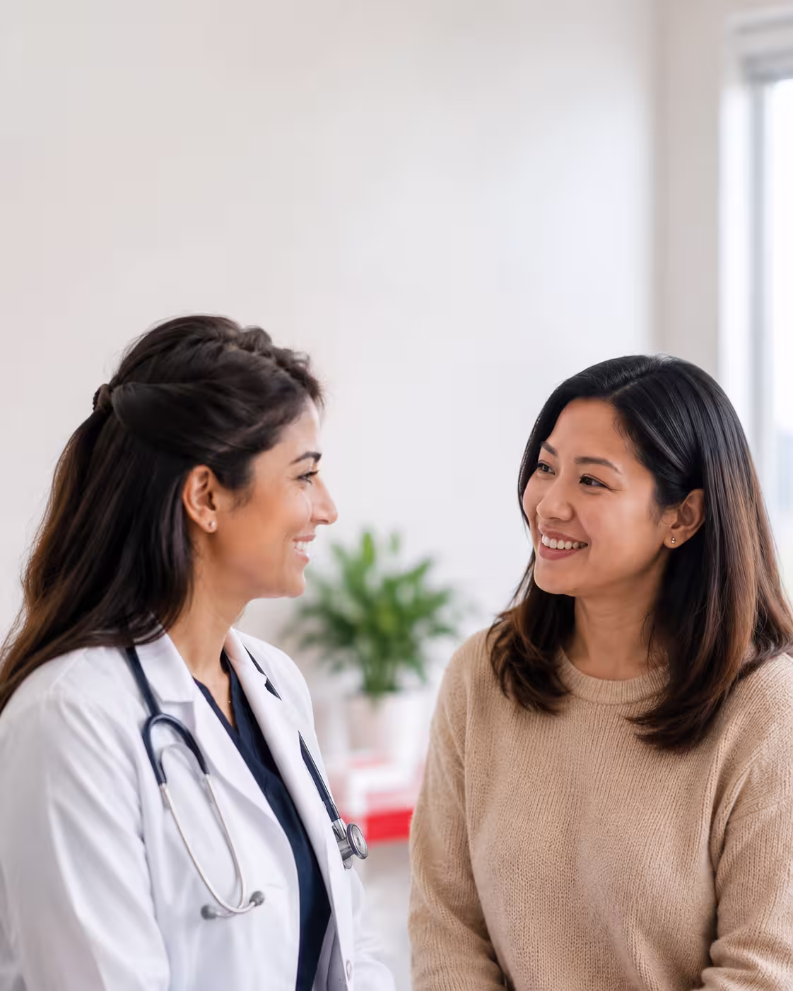 friendly female physician in a white coat consulting with a smiling adult patient in a bright, modern medical clinic exam room.
